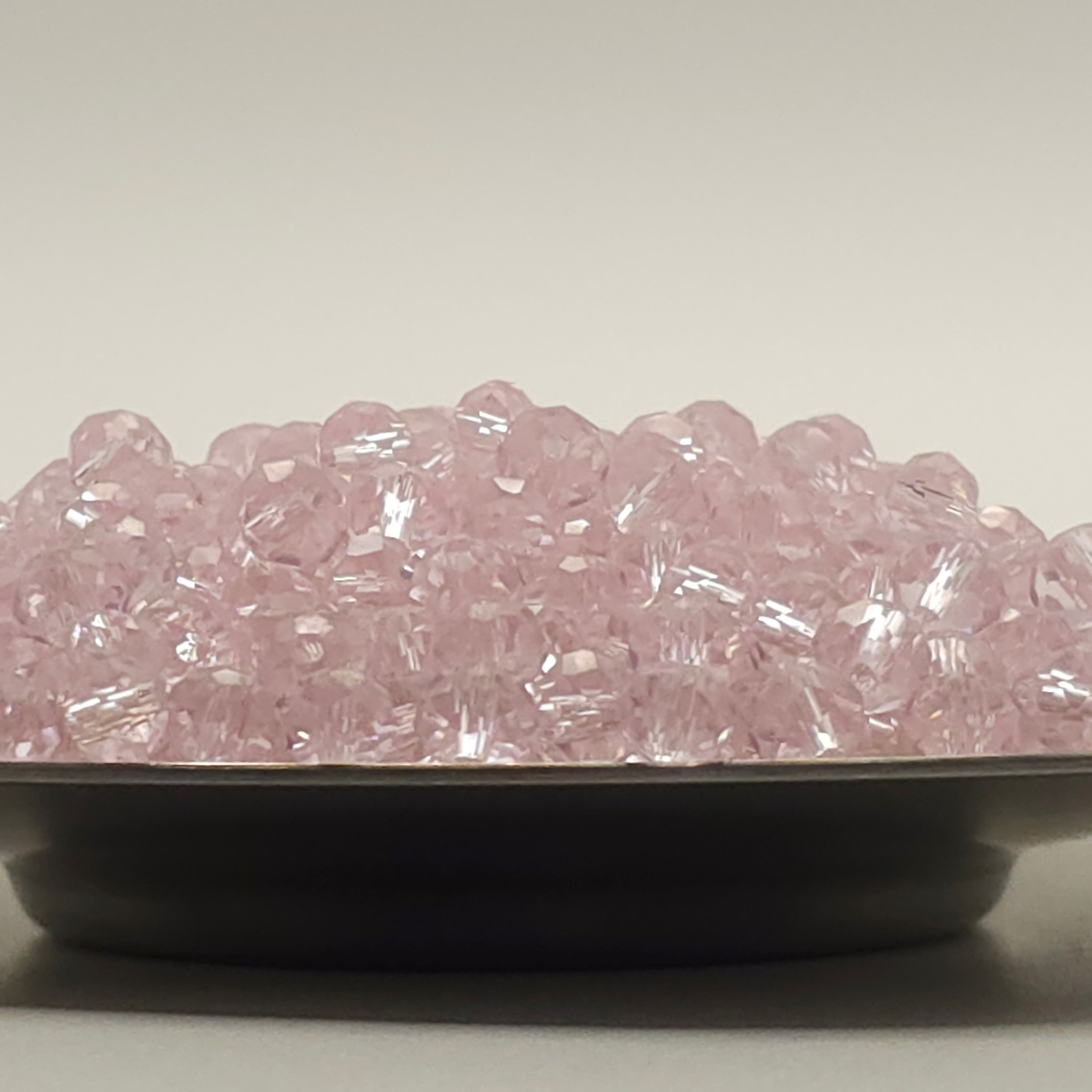 Pink faceted glass beads piled in a shallow black dish against a plain light gray background.