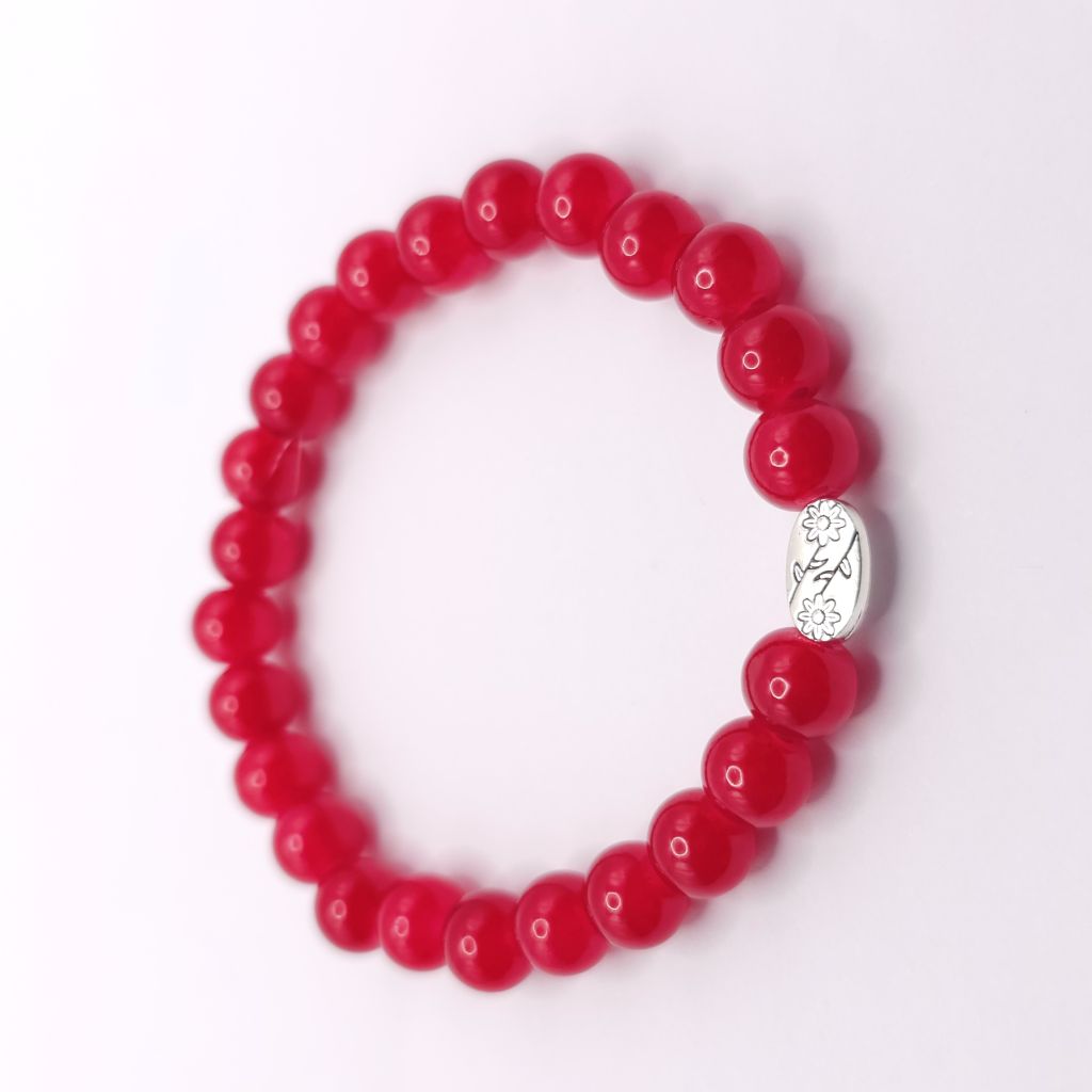 A red beaded bracelet with a small silver oval charm featuring a floral pattern, photographed on a clean white background.