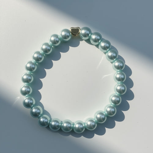 A beaded bracelet made of glossy, aquamarine green pearlised glass beads with a small gold heart-shaped charm, arranged in a circle on a white surface with soft shadows.