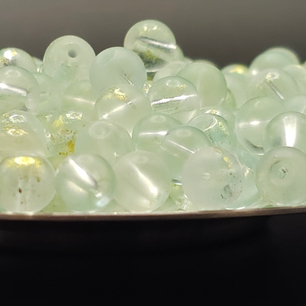 A close-up pile of small, round, translucent pale green beads with holes, resting on a dark surface.