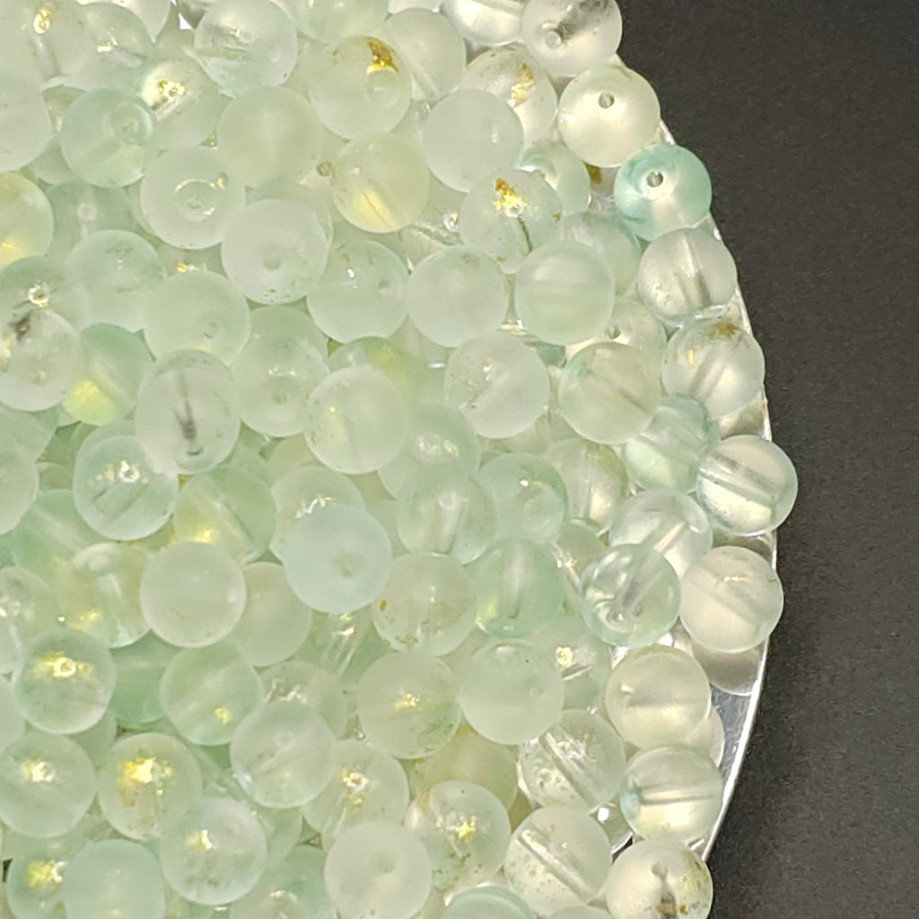 A close-up of many small round translucent beads in a pale mint green and milky white tone, piled on a dark surface with a metallic edge visible on the right.
