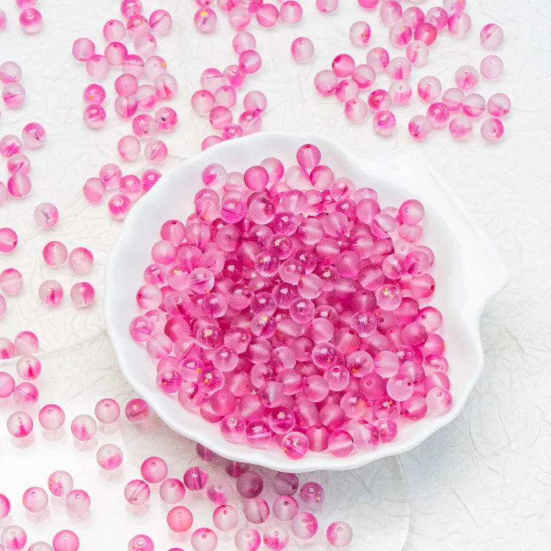 Pink translucent glass beads scattered on a white textured surface, with a small white dish filled with the beads in the center.