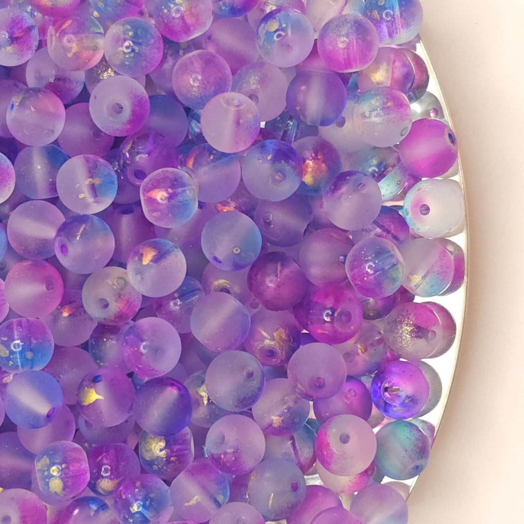 A close-up pile of round translucent beads in shades of purple, lavender, and blue with iridescent glitter, shown on a light beige background.