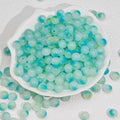Light blue and mint green marbled acrylic beads with hollow centers, displayed in a white scalloped dish with some beads scattered around on a textured white background.