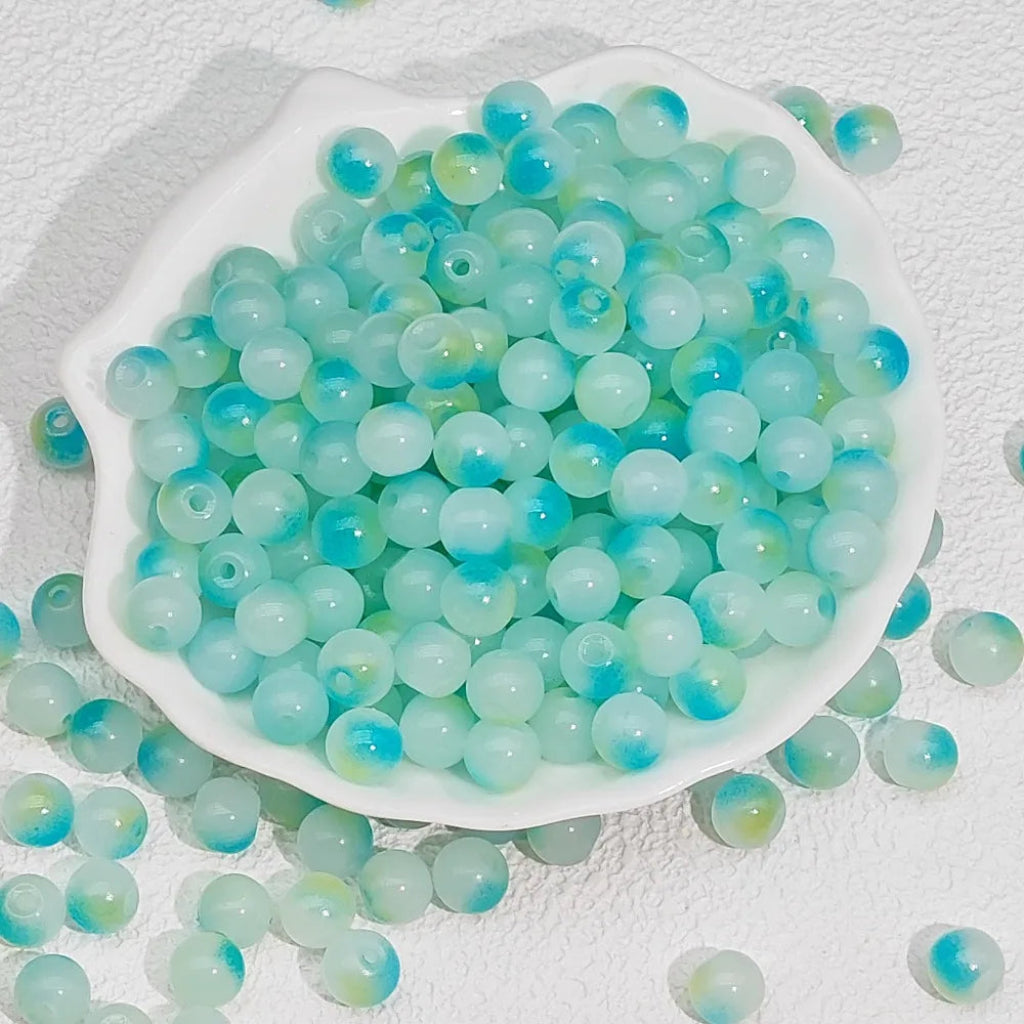 Light blue and mint green marbled acrylic beads with hollow centers, displayed in a white scalloped dish with some beads scattered around on a textured white background.