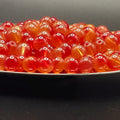 A close-up of many small translucent red-orange beads piled in a shallow dish against a dark background.
