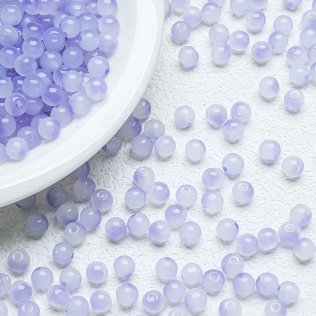 Light purple translucent round beads scattered on a white textured surface, with a white bowl filled with more beads in the upper left corner.