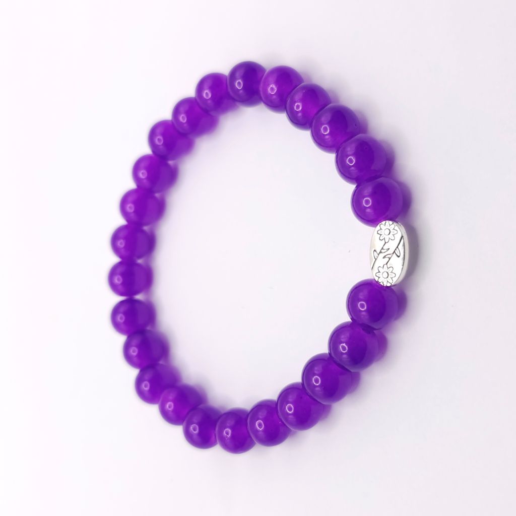 A purple beaded stretch bracelet with a small silver oval charm featuring a floral design, displayed on a clean white background.
