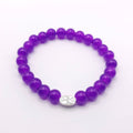 A purple beaded stretch bracelet with a small silver oval charm featuring a floral design, displayed on a clean white background.