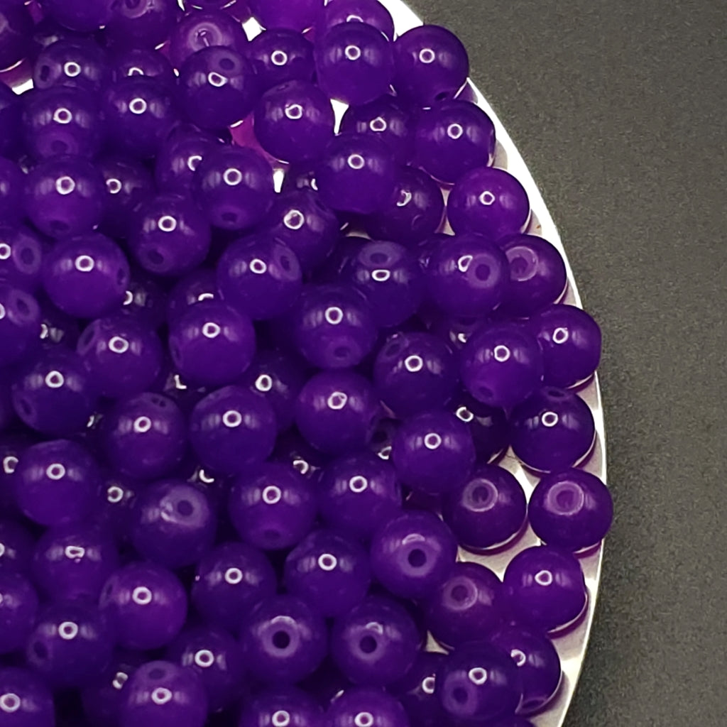 A close-up of many glossy purple round beads with central holes, piled on a white plate against a dark gray background.