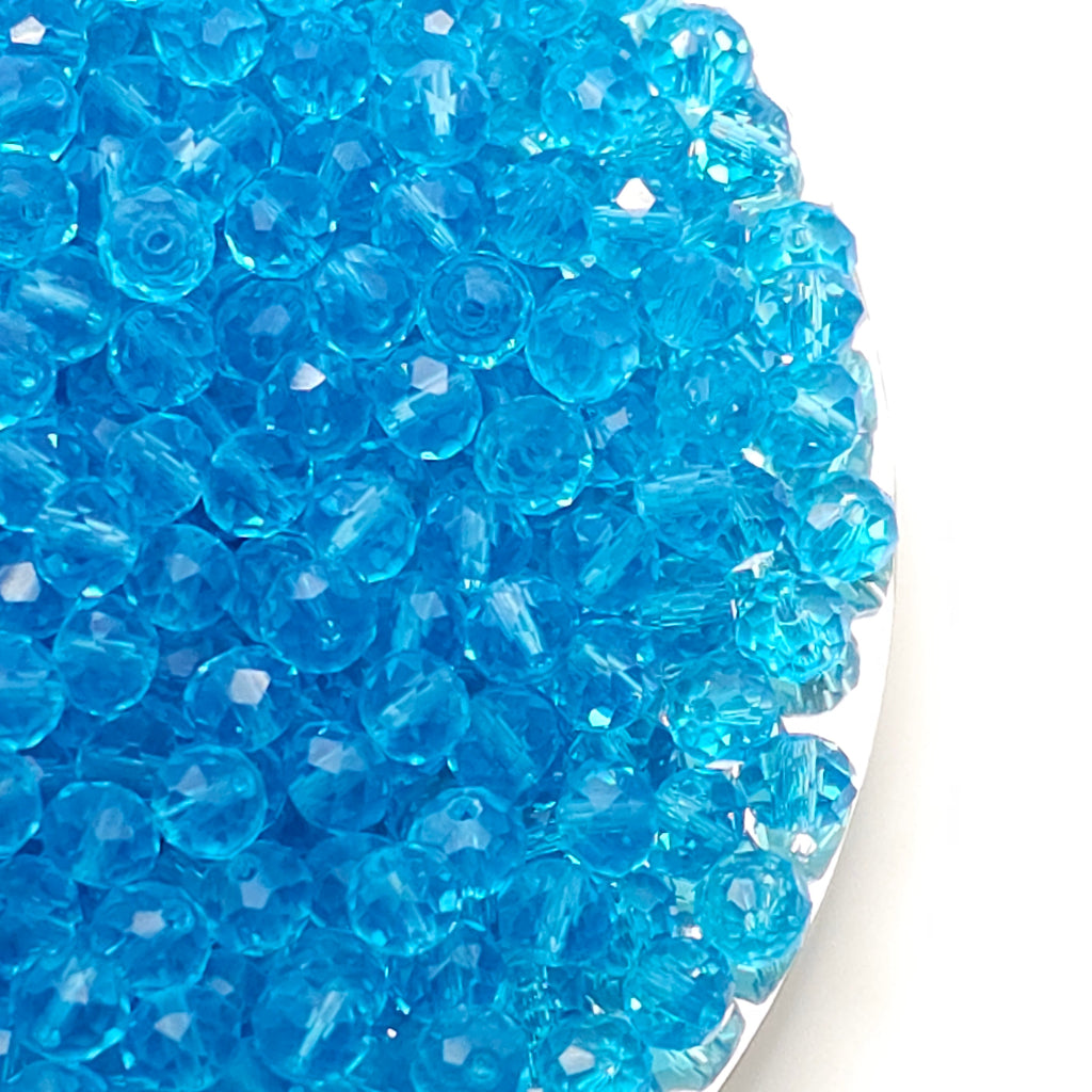 A close-up of many small translucent light blue faceted beads piled on a white surface.