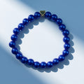 A beaded bracelet made of glossy deep blue pearlised beads with a small gold heart-shaped charm, arranged in a circle on a light blue background with soft shadows.