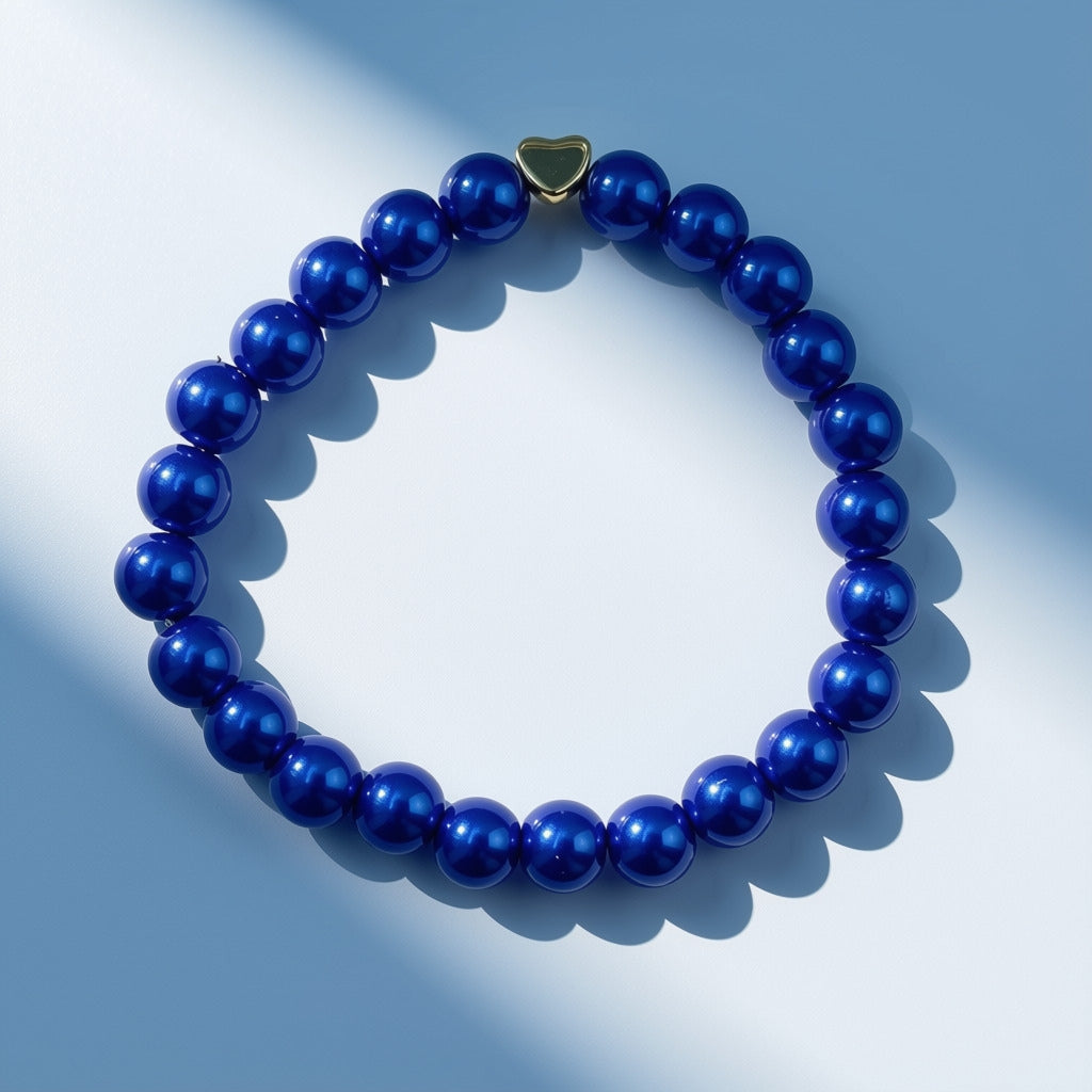 A beaded bracelet made of glossy deep blue pearlised beads with a small gold heart-shaped charm, arranged in a circle on a light blue background with soft shadows.