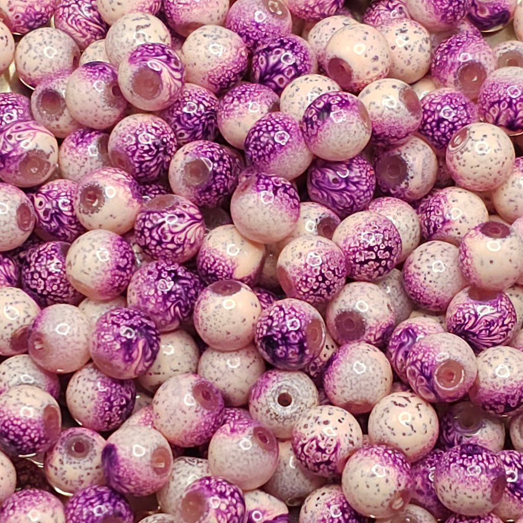 A close-up of many round glass beads in a silver bowl, featuring a marbled pattern of purple, pink, and white.