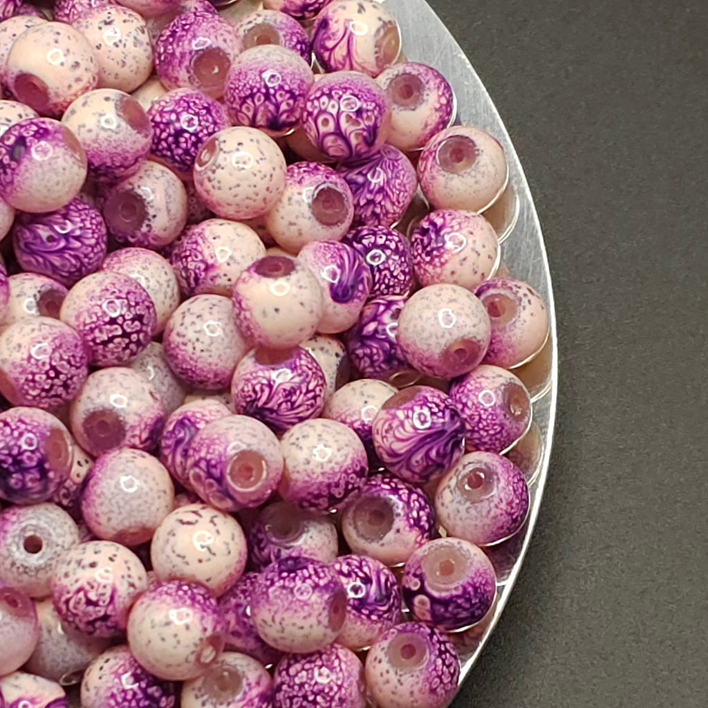 A close-up of many round glass beads in a silver bowl, featuring a marbled pattern of purple, pink, and white.