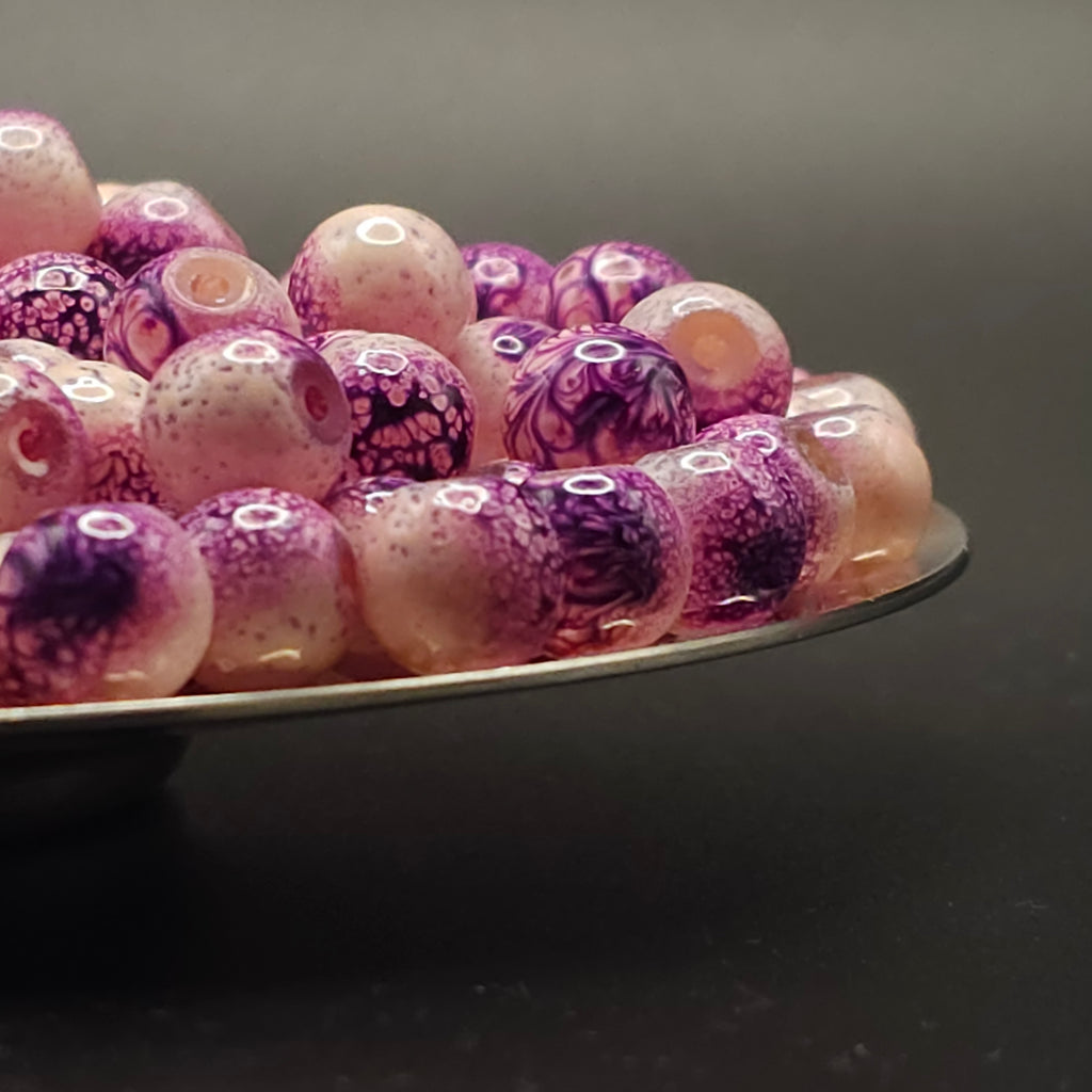 A pile of round glass lampwork beads in shades of pink, purple, and white with marbled patterns, displayed on a silver metal tray against a dark gray background.
