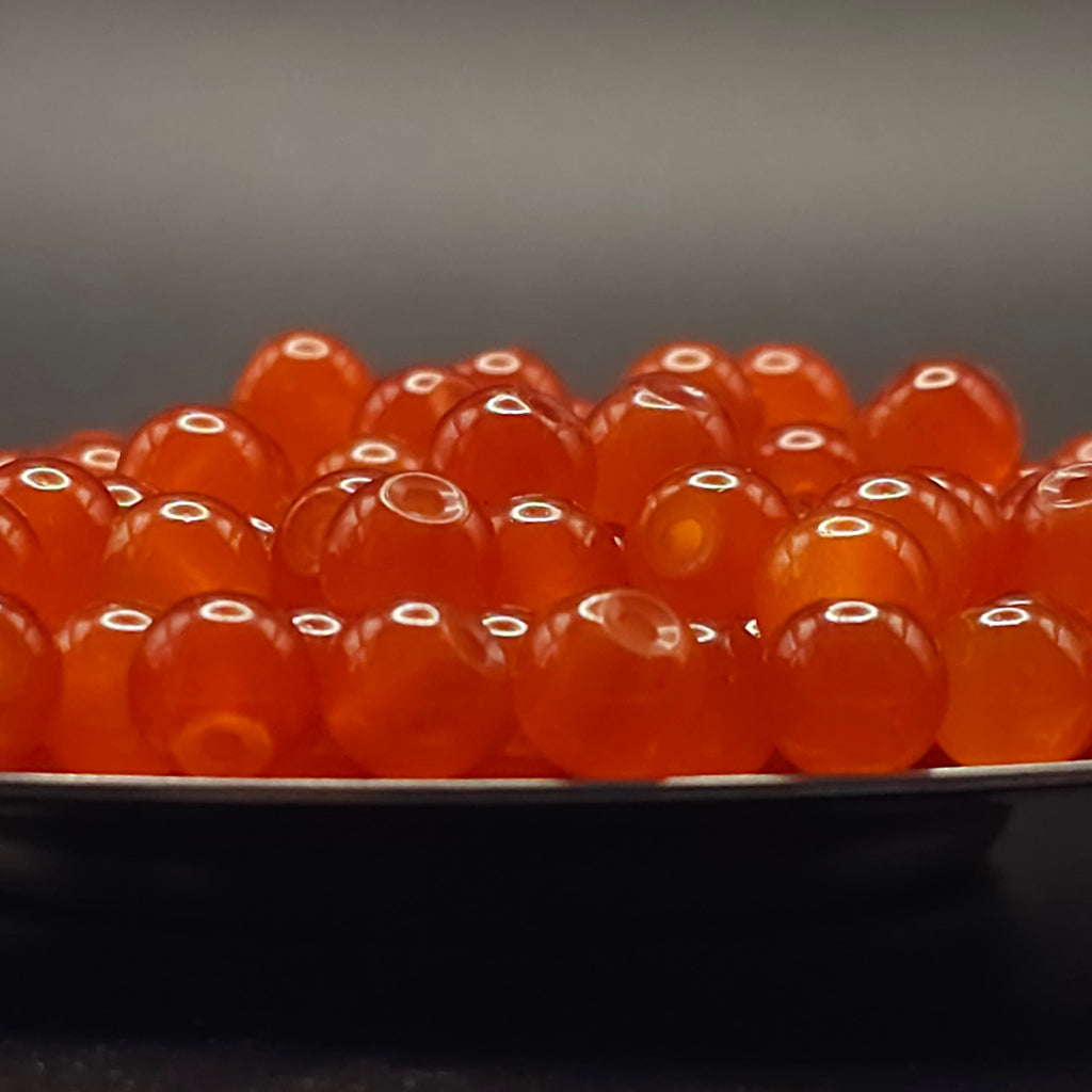 A close-up of glossy translucent orange-red round beads or gemstones piled on a dark surface against a dark gray background.
