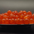 A close-up of glossy translucent orange-red round beads or gemstones piled on a dark surface against a dark gray background.