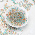 Small round translucent beads in aqua blue, white, and peach-orange tones, with some scattered around and others piled in a white scalloped dish on a light textured background.