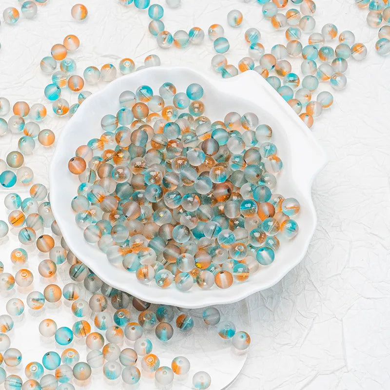 Small round translucent beads in aqua blue, white, and peach-orange tones, with some scattered around and others piled in a white scalloped dish on a light textured background.