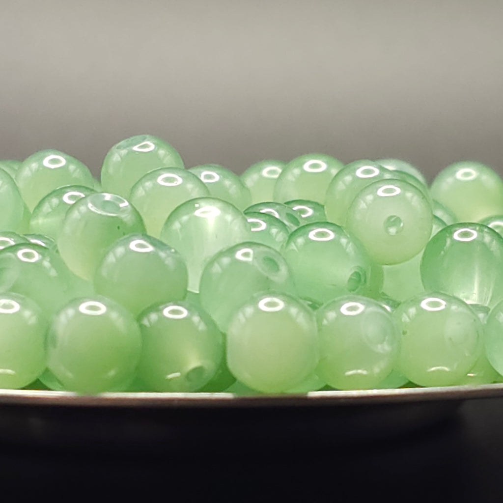 A close-up of round, pale green translucent beads or gemstones piled in a shallow dish against a soft gray background.