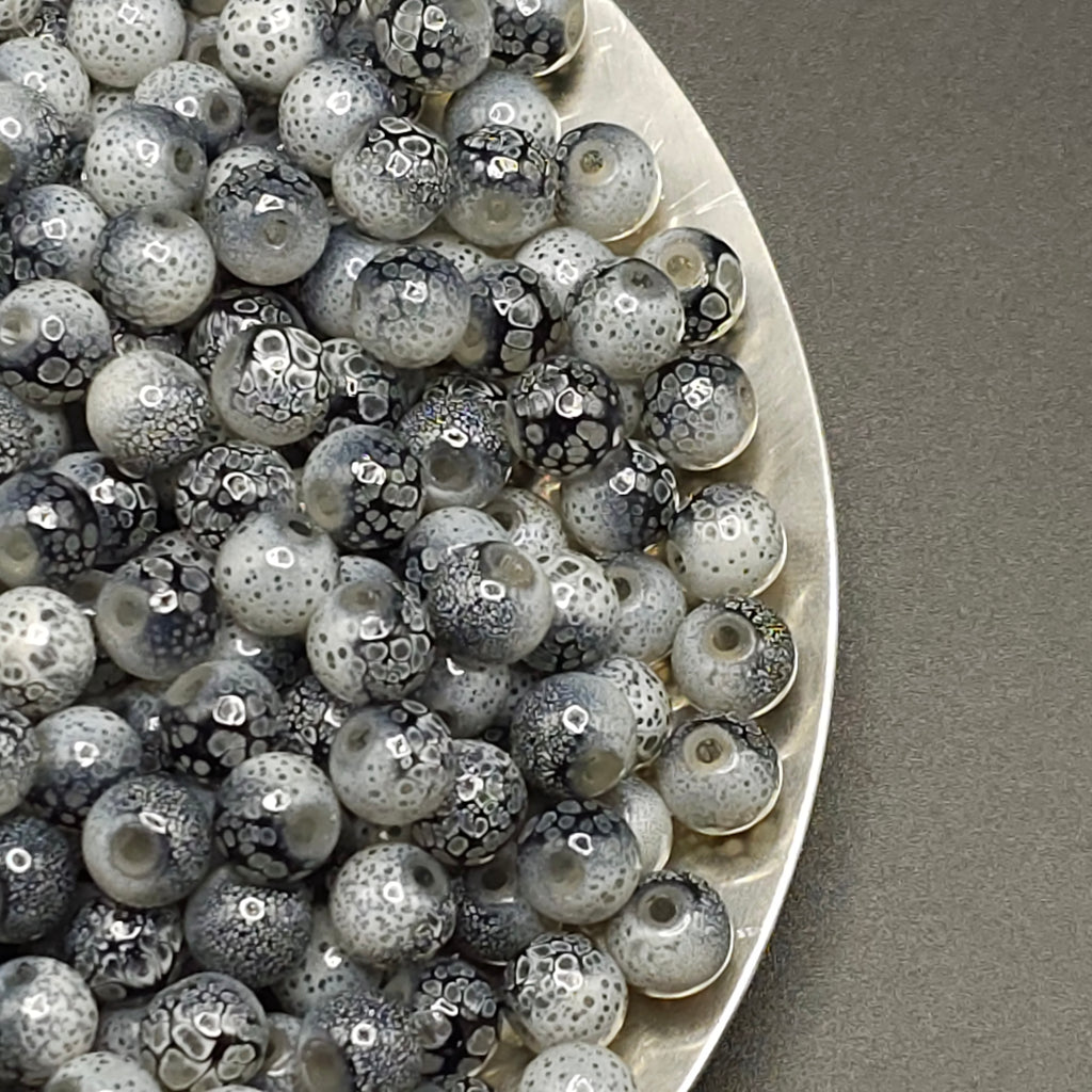 A close-up of many round glass beads with a mottled black, white, and gray spotted pattern, piled on a white plate against a gray background.