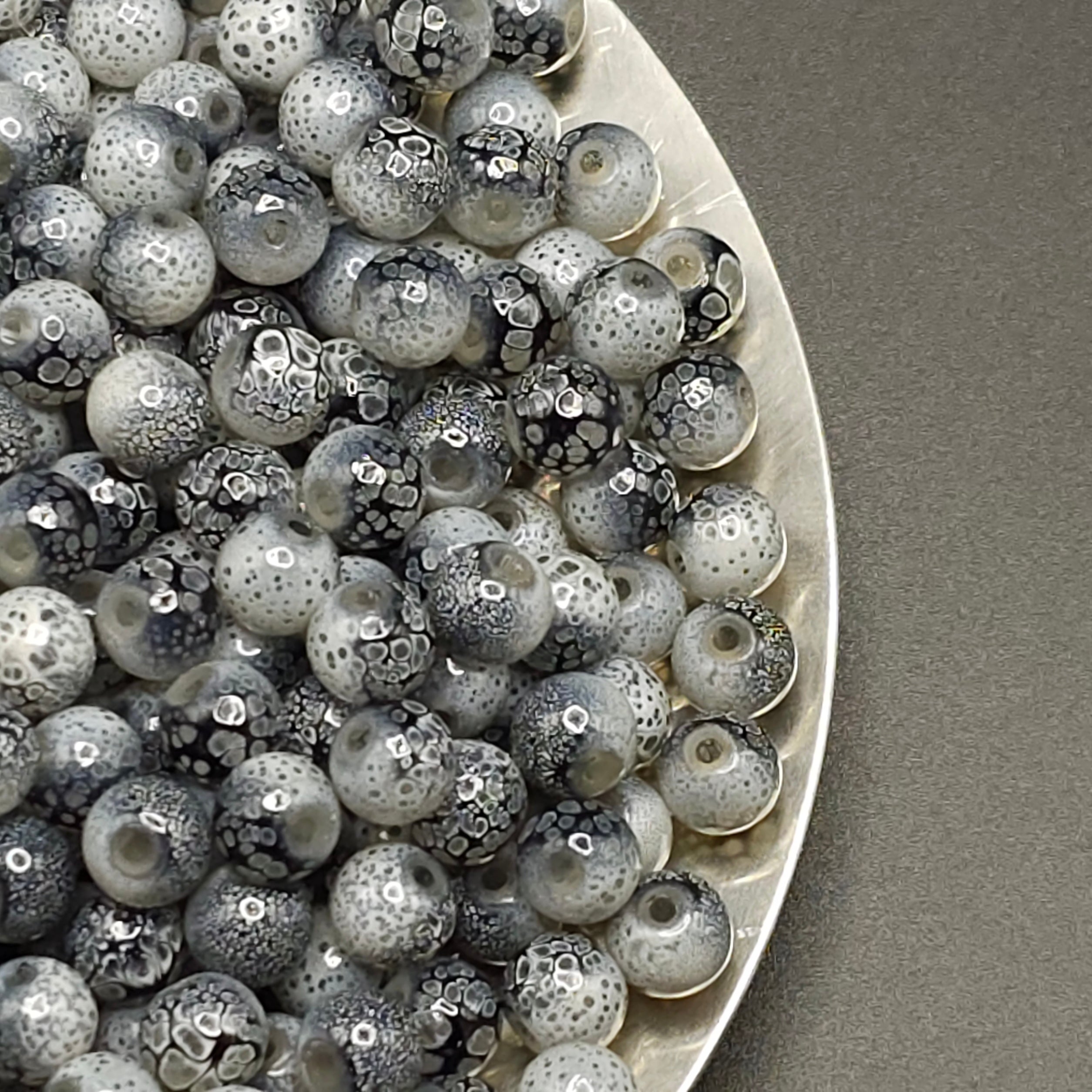 A close-up of many round glass beads with a mottled black, white, and gray spotted pattern, piled on a white plate against a gray background.