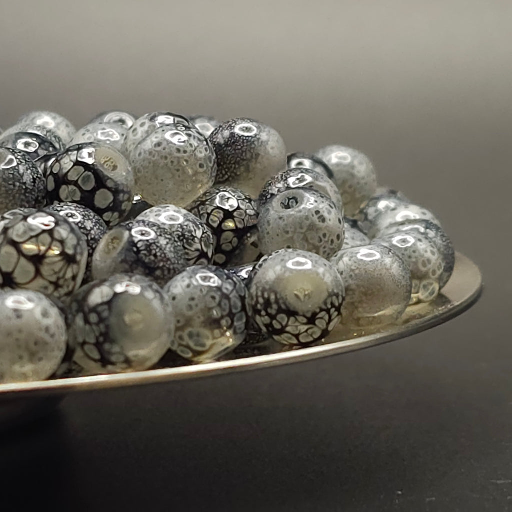 A pile of round glass beads with a black and white spotted pattern, resting on a silver metal tray against a dark gray background.
