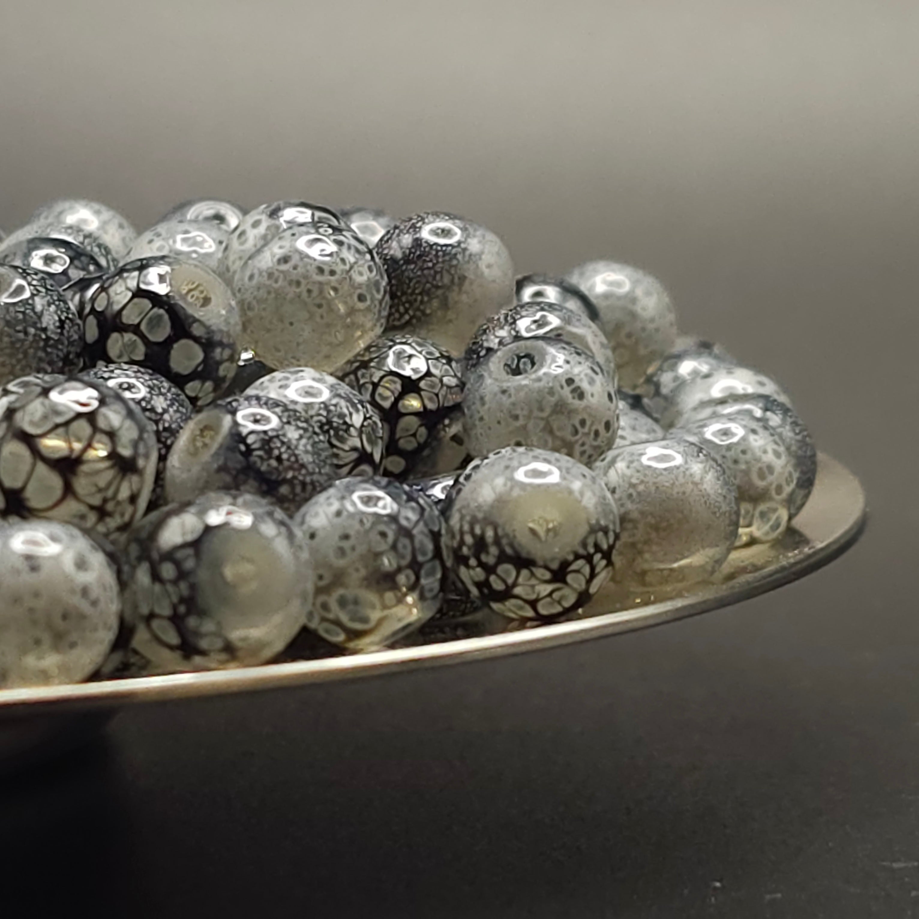 A pile of round glass beads with a black and white spotted pattern, resting on a silver metal tray against a dark gray background.