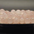 A close-up of multiple pale pink translucent oval beads arranged in rows on a dark surface against a soft gray background.