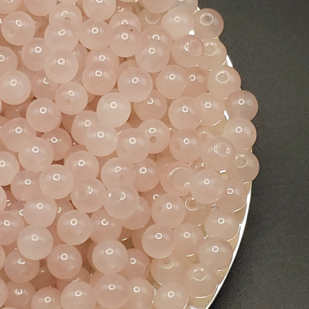 A close-up of many small round translucent beads in a pale blush pink color, piled on a dark gray surface with part of a silver container edge visible.