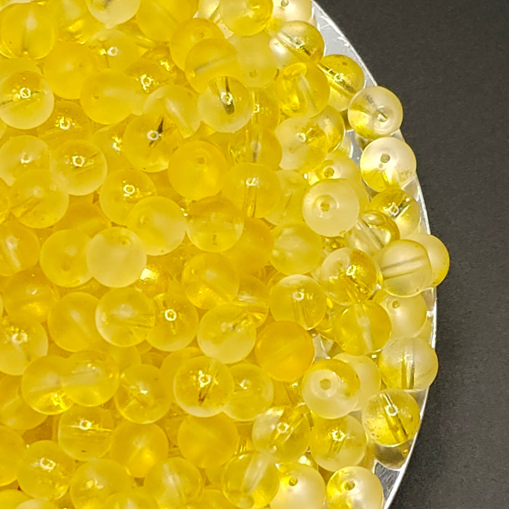 A pile of small, round, translucent yellow beads with a double-hole design, resting on a silver tray against a dark gray background.