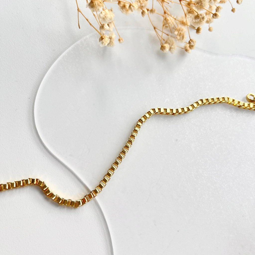 Gold chain anklet on a white surface with dried flowers.