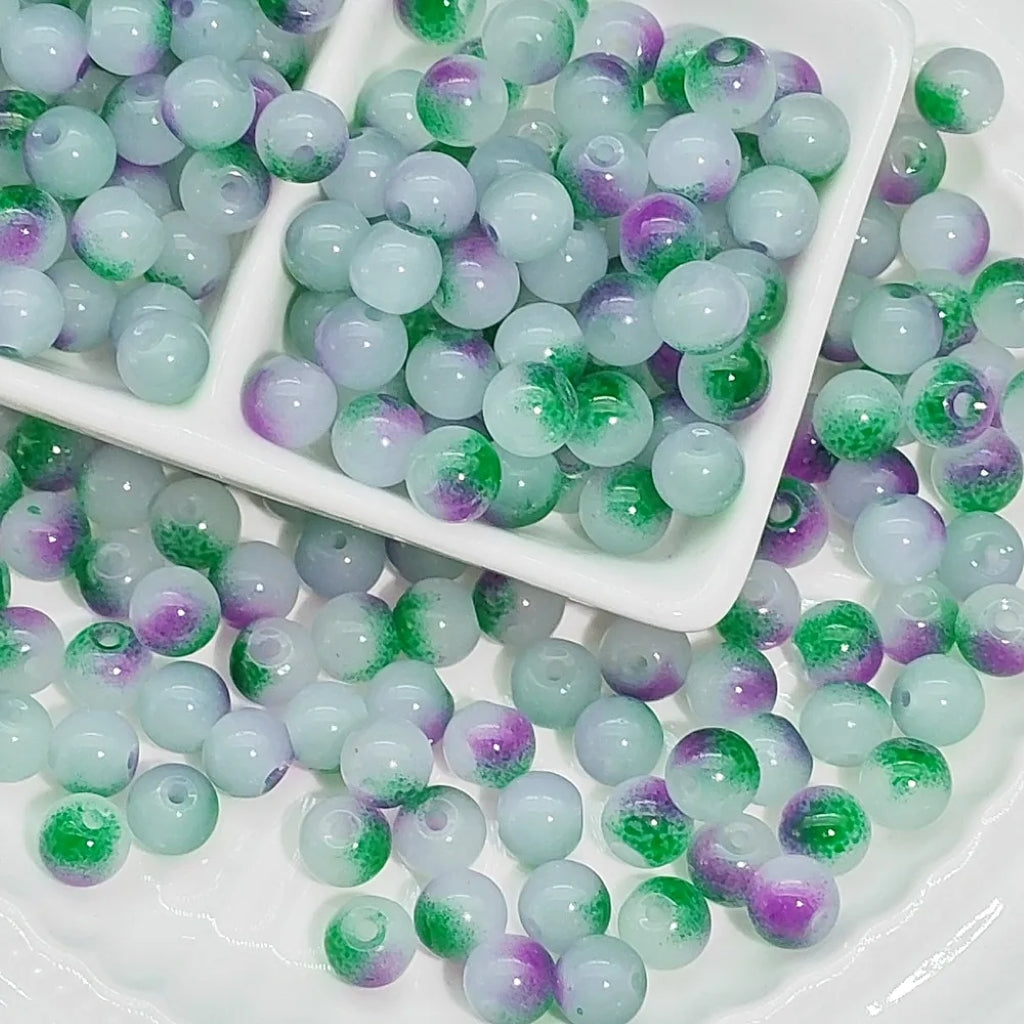 Round glass beads with a light aqua-blue base, featuring green and purple glittery swirls, displayed on a white plate with a small white divided dish holding some of the beads.