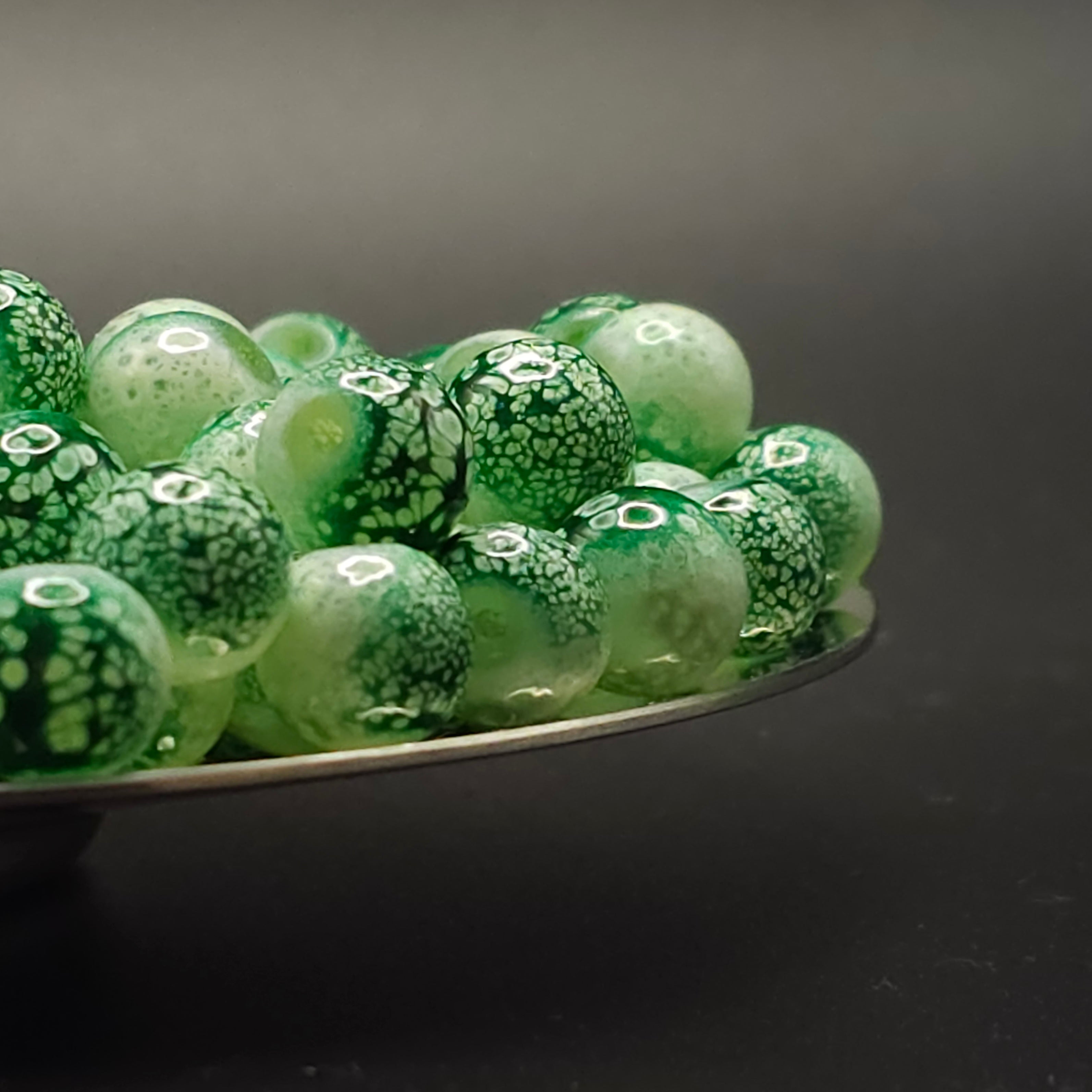 A pile of round glass beads in shades of green with white marbled or cracked patterns, displayed on a dark reflective surface against a dark gray background.