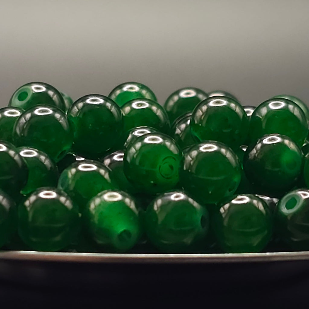 A pile of translucent green glass beads with hollow centers, resting in a dark container against a neutral gray background.