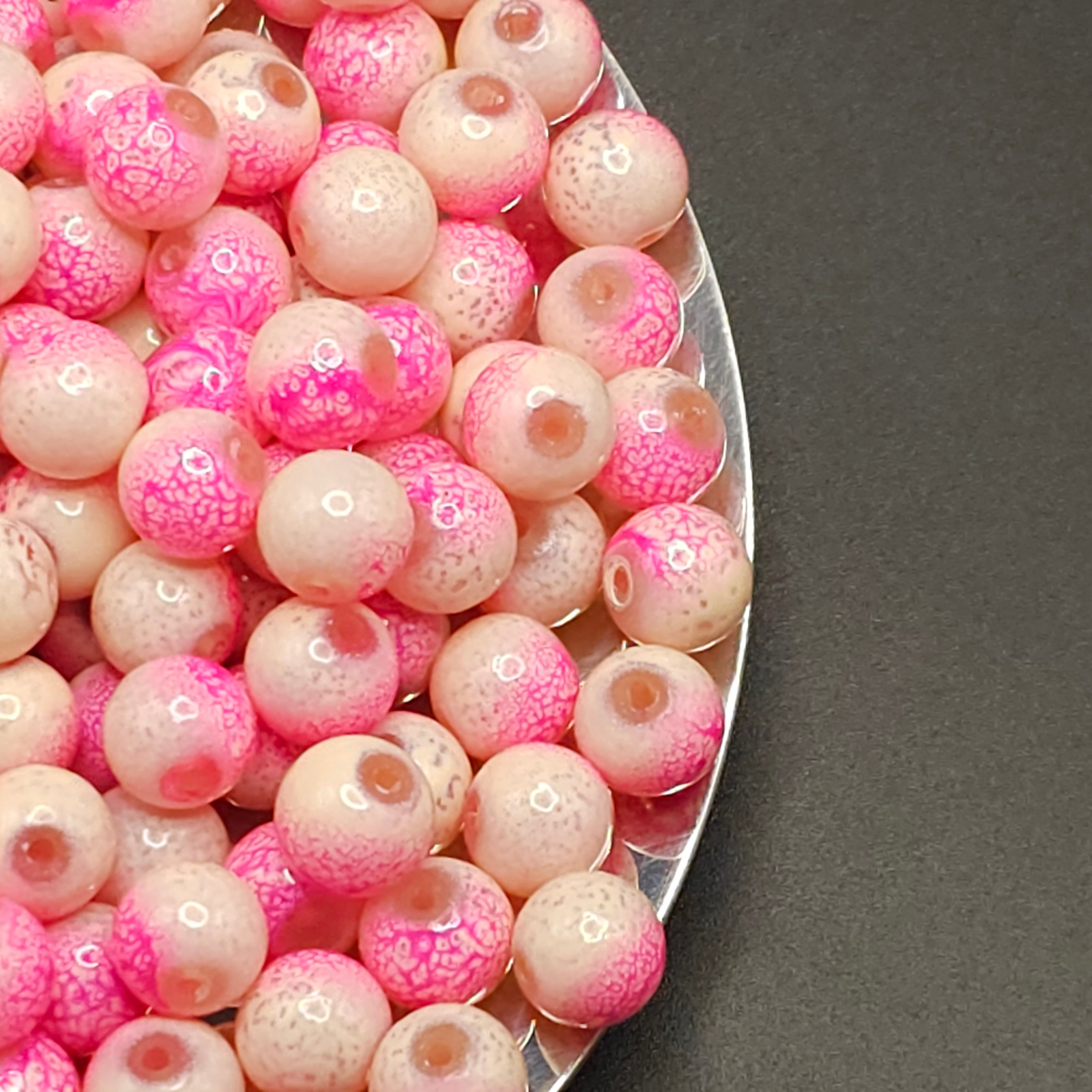 A close-up of many round glass beads in pink and pale peach tones, some with bright pink mottled patterns and others with a glossy pink center, arranged on a silver tray against a dark gray background.