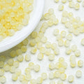 Small translucent pale yellow round beads scattered on a white textured surface, with a white plate partially visible at the top left containing more beads.