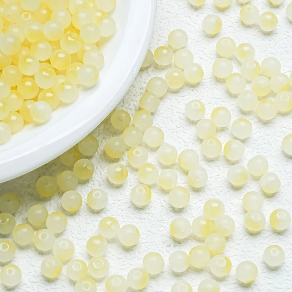 Small translucent pale yellow round beads scattered on a white textured surface, with a white plate partially visible at the top left containing more beads.
