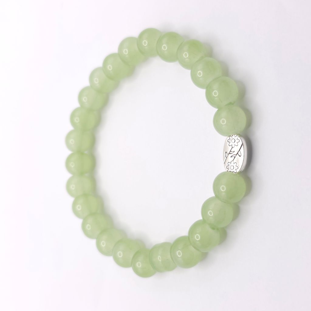 A light green beaded stretch bracelet with a small silver oval charm featuring a floral engraving, displayed on a clean white background.