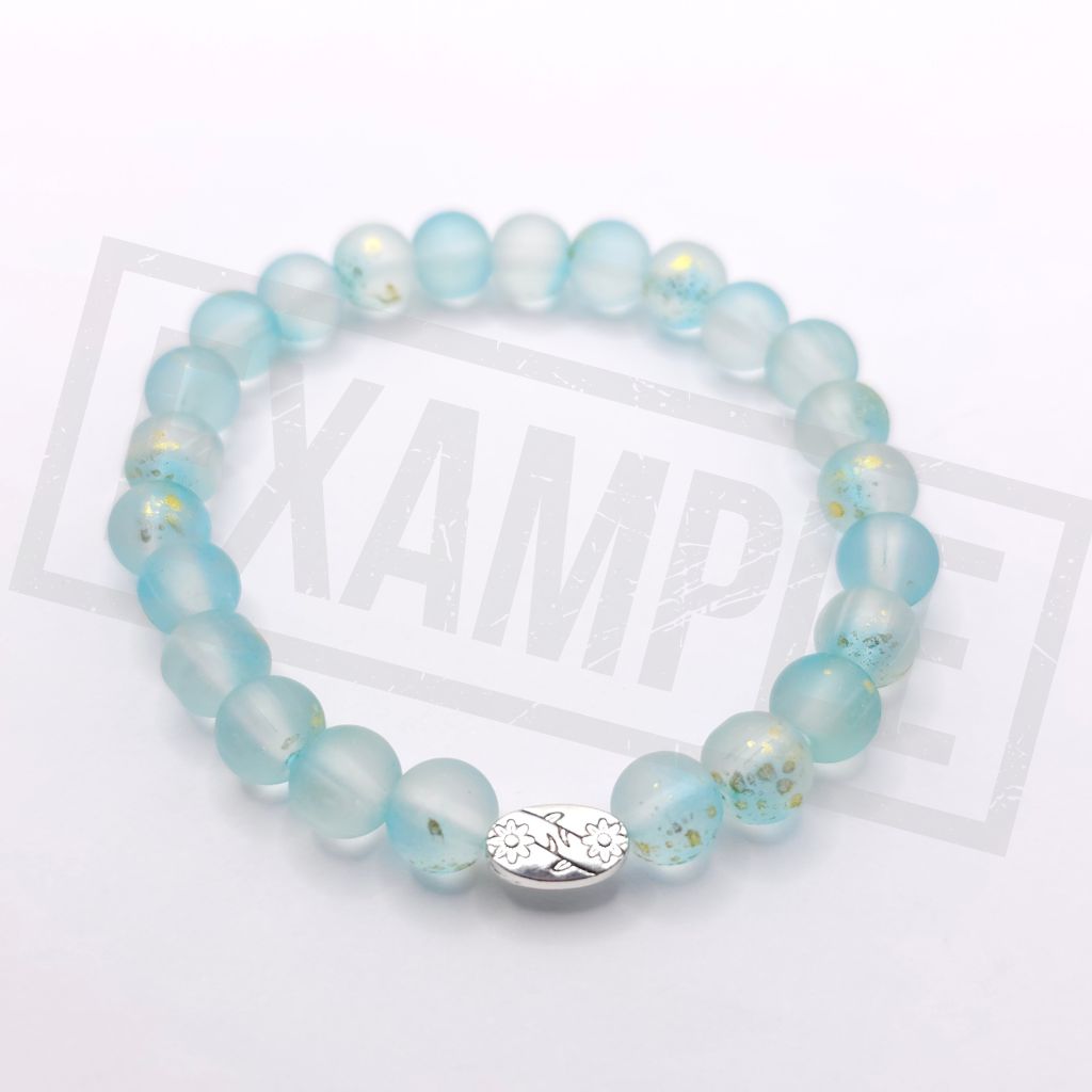 A beaded stretch bracelet made of light blue translucent round beads with small gold flecks, featuring a silver-toned metal charm with a floral design and the letters 'JM'. The bracelet is displayed on a white background.