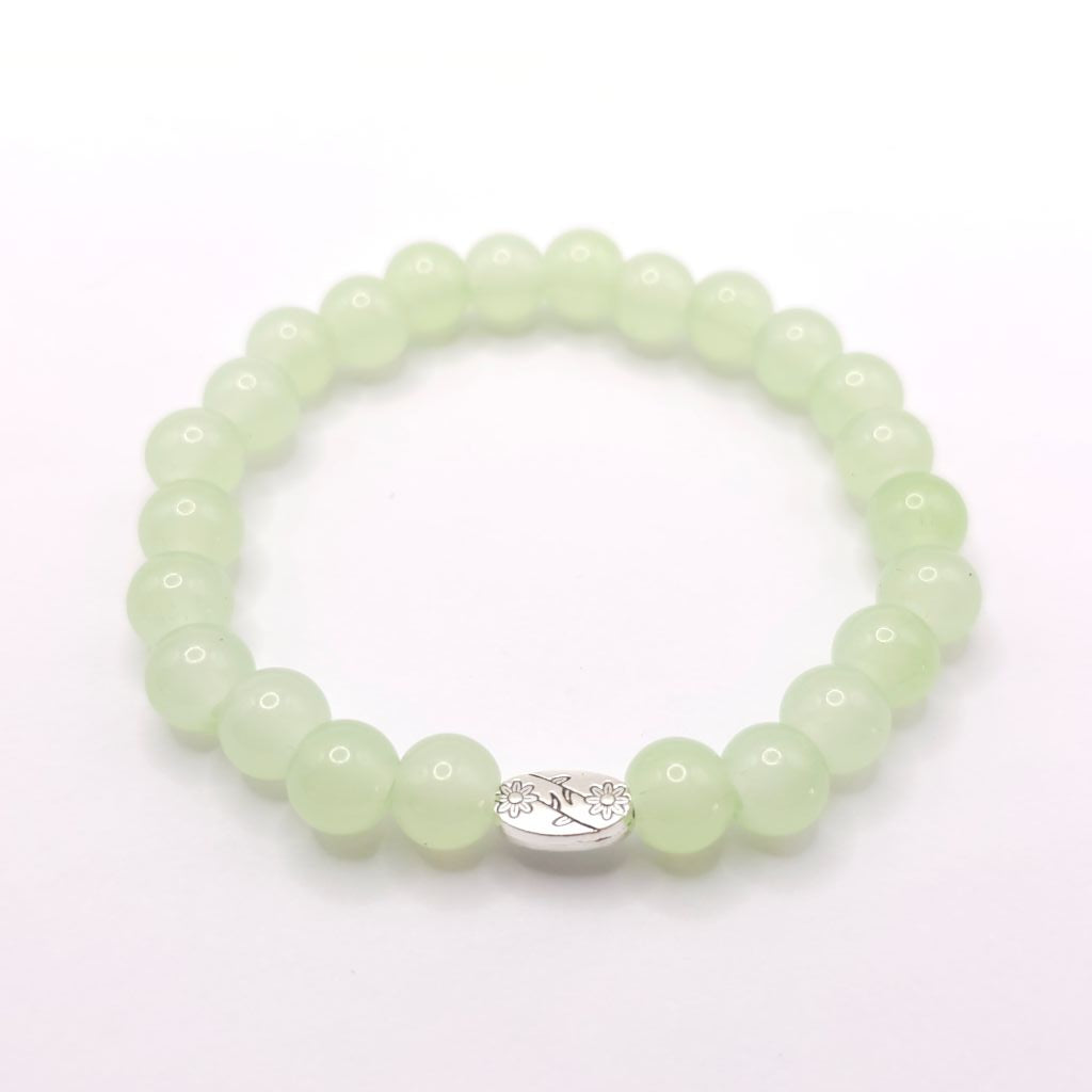 A beaded stretch bracelet made of light green translucent round beads with a small silver-toned oval charm featuring a floral design, shown on a plain white background.