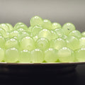 A pile of round, translucent pale green beads with holes, displayed on a dark surface against a neutral gray background.