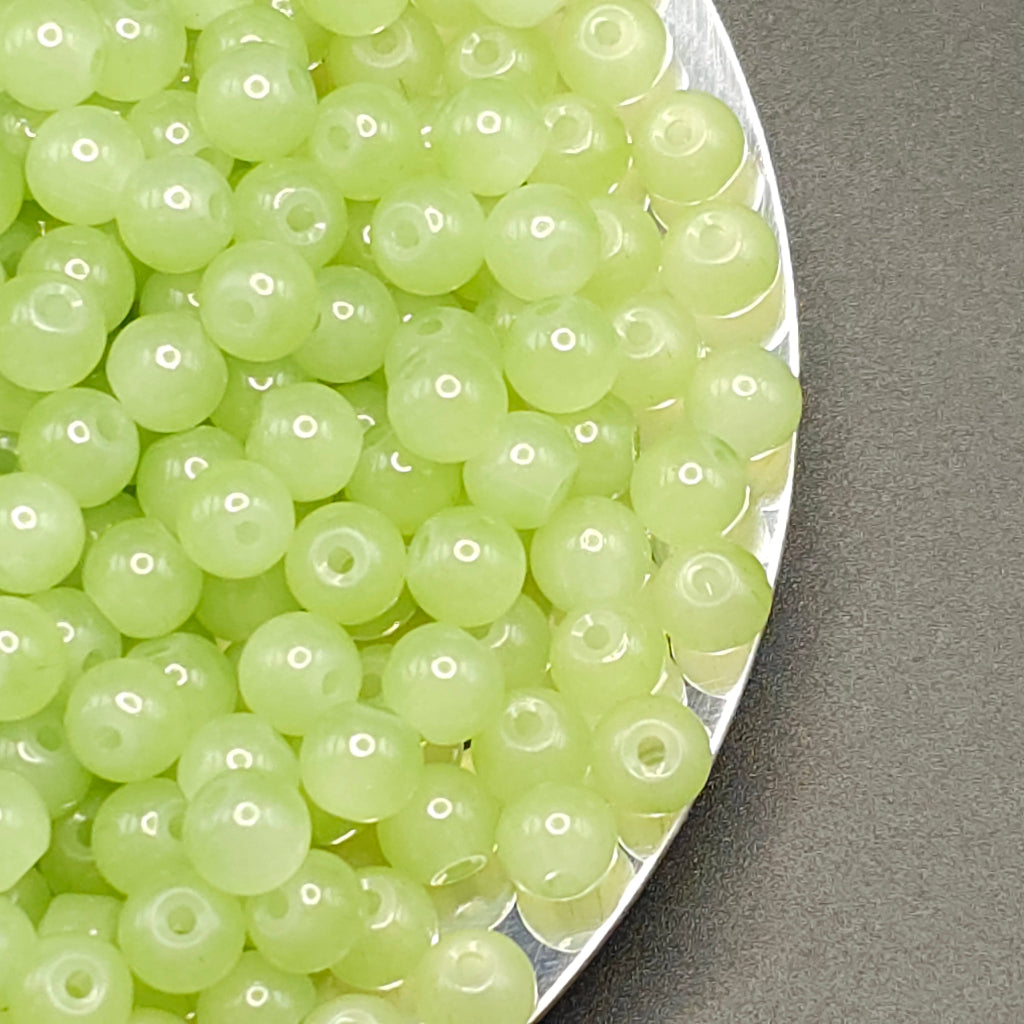 A close-up of many small translucent pale green round beads with center holes, piled on a silver tray against a gray background.
