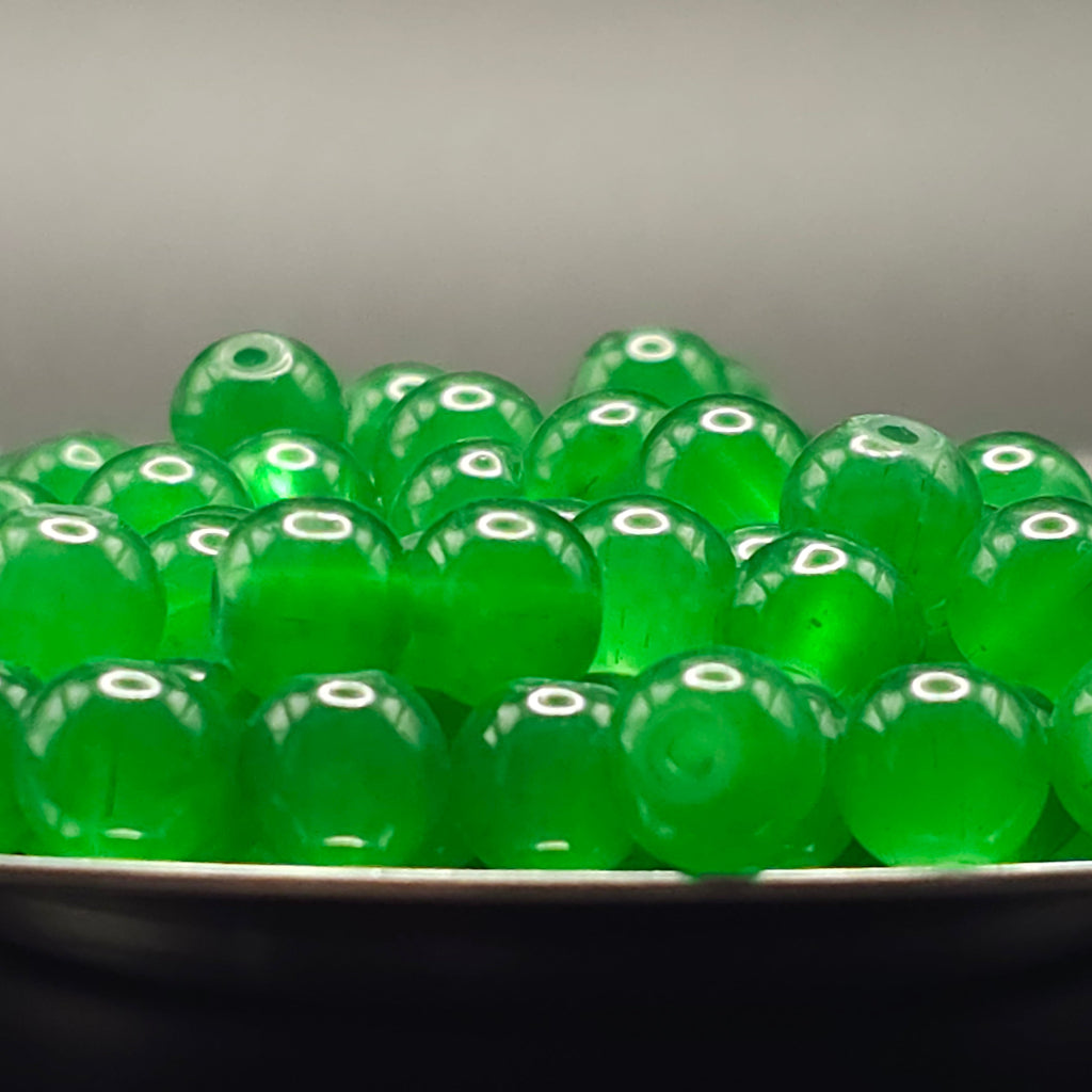 A pile of translucent green glass beads with hollow centers, resting on a dark surface against a neutral gray background.