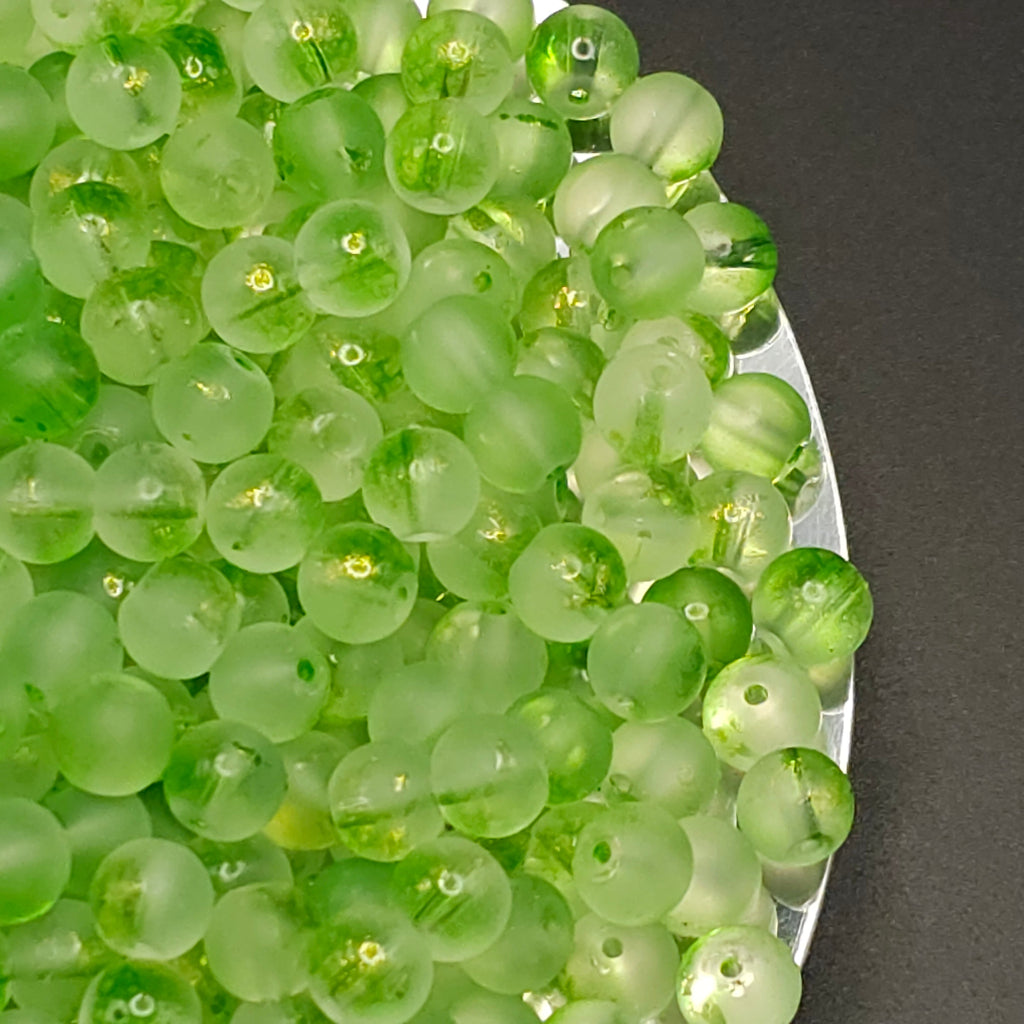 A pile of round, translucent light green beads with small holes, shown close-up on a dark surface next to a silver container.