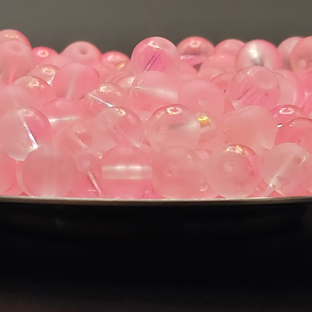 A close-up of many translucent pink round beads or capsules piled together in a shallow dish, with a dark background.