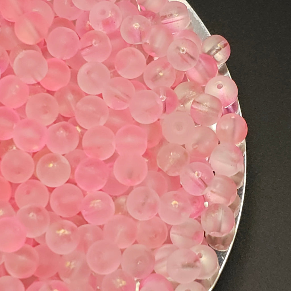 A close-up of many small translucent pink round beads piled on a silver tray against a dark background.