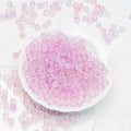 Pink translucent round craft beads scattered on a white textured surface, with a white scalloped dish filled with the beads in the center.