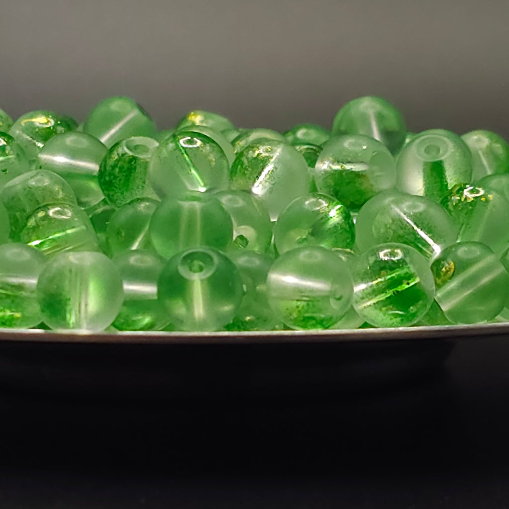 A close-up of translucent green glass beads with internal green inclusions, piled on a dark surface.